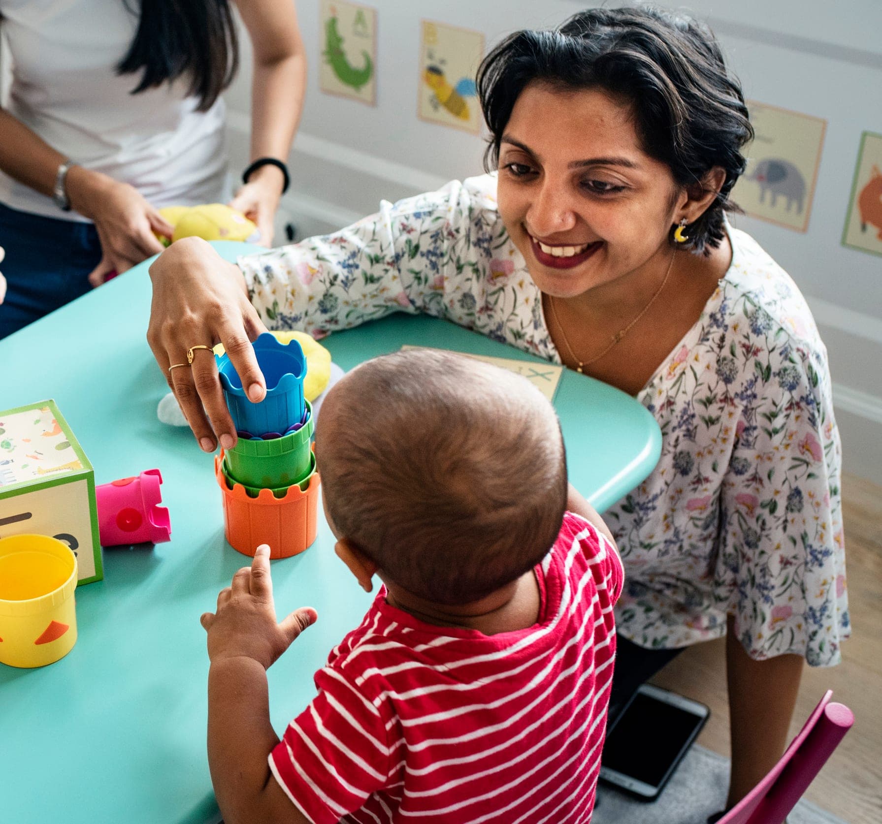 A Wee Discover teacher smiling with a toddler stacking colorful cups at a classroom table