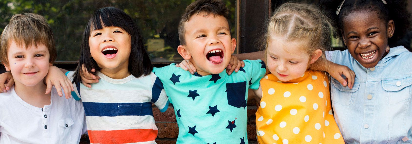 Children smiling and laughing sitting on a brick wall outside in colorful clothing.